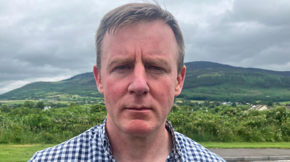 A man with brown hair, blue and white check shirt and mountains in the background