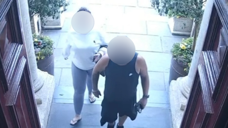 A woman in a white sweatshirt and leggings and a man in a black vest and black shorts walking into a building with red doors. Both of their faces are blurred.