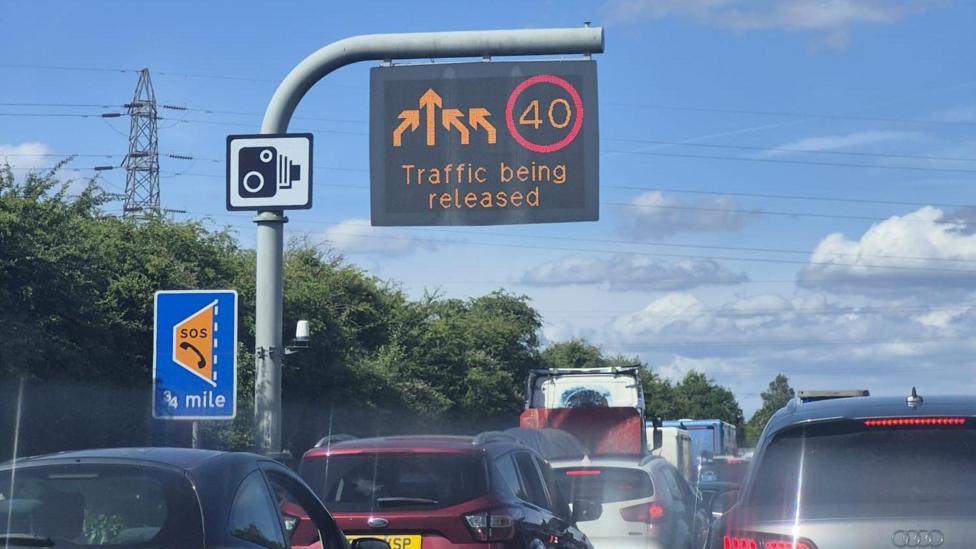 Traffic queues on the motorway. A sign with the text 'Traffic being released' as well as arrows and a temporary speed limit of 40 MPH hangs above.