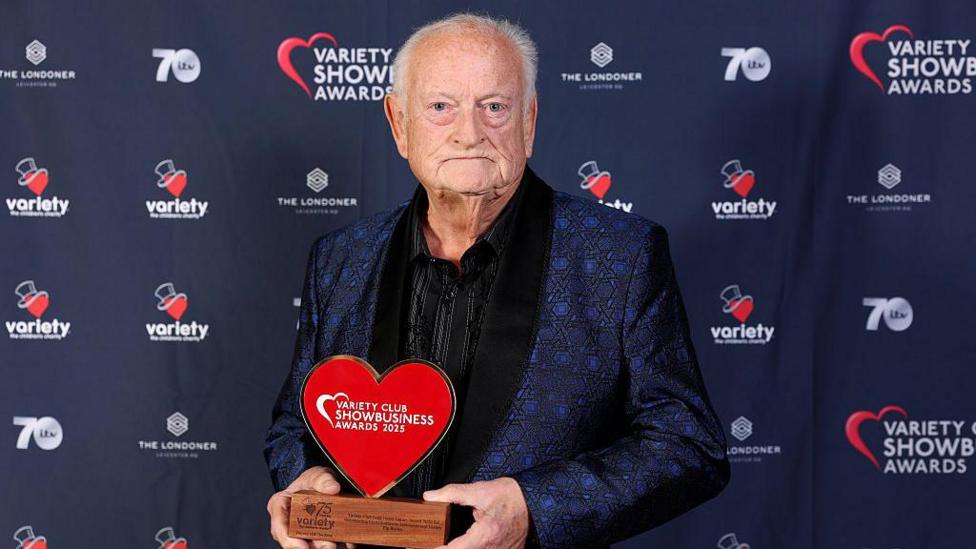 Pip Burley poses with a trophy at the Variety club Gold Heart Legacy Award 2025. He wears a navy patterned suit, and black shirt. He has short grey hair and is bald on the top of his head.