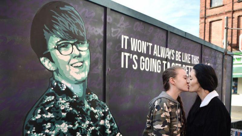 Robyn Eswards-Peoples (left) is wearing a camouflage jacket and grey hoodie. Sharni Edwards-Peoples (right) is wearing a black coat. They are kissing in front of the Lyra McKee mural in Belfast. Lyra is seen smiling and is wearing a colourful shirt. She is beside a quote reading: It won't always be like this. It's going to get better'