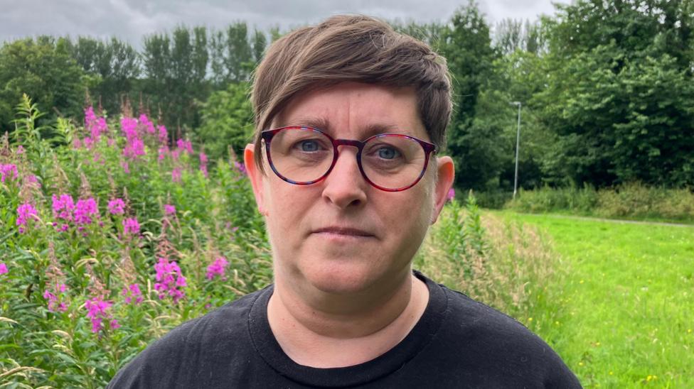 Cali Morrow has short brown hair and is wearing pink glasses and a back shirt. She is standing in a large grassy field with flowers.