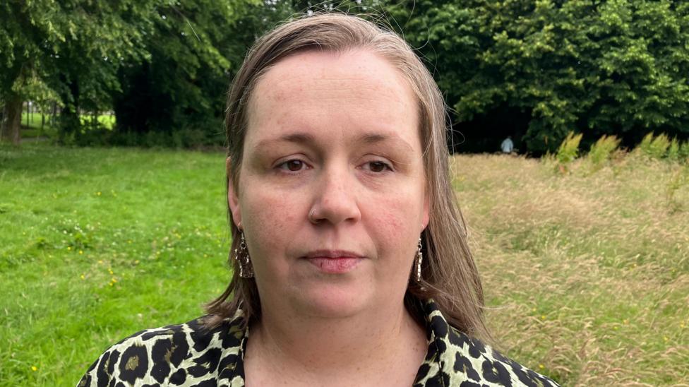 Julie McCloskey is standing in a large field with a number of purple plants and trees. She is wearing a leopard print top and has short brown hair.