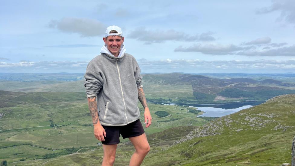 Ed Shirt wearing a hoodie and shorts standing on a mountain with a view behind him