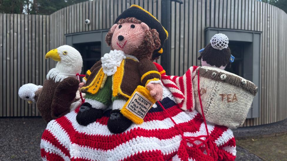 An assortment of knitted figures sits on top of a post box. A crocheted American flag lies underneath a knitted eagle, a little figure wearing old style officer clothing and a naval hat and a box of tea. It's all brightly coloured and striking. 