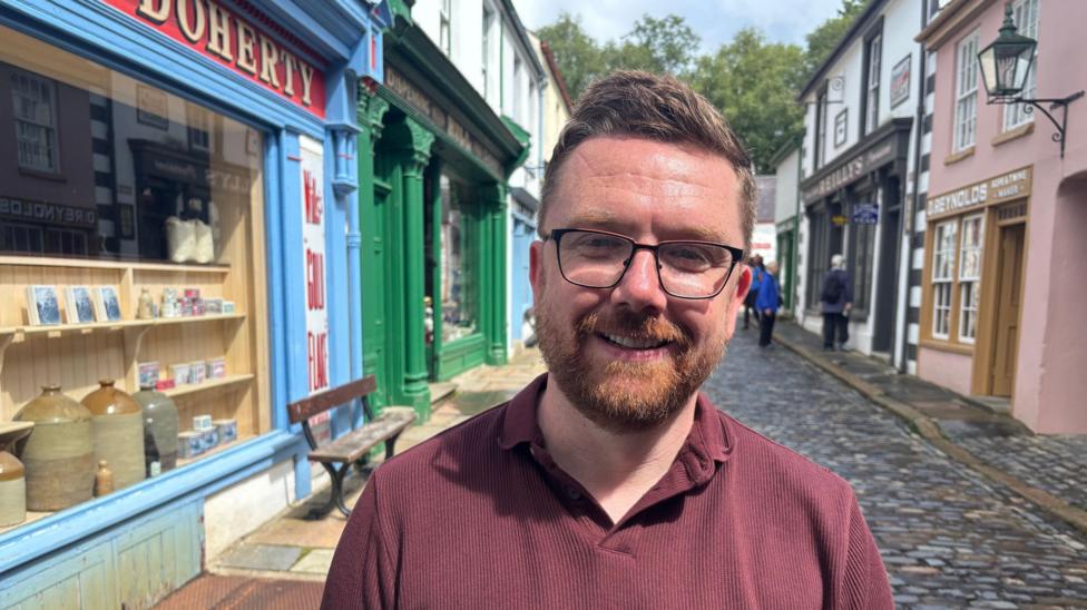 A man stands on a quaint street. He is wearing a maroon polo shirt and glasses. He has quaffed brown hair and sports a red beard. The street behind him is cobblestoned with ole style shops on either side.