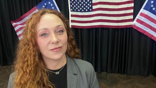 A woman stands in front of a display of 3 American flags. She has long red curly hair that hangs over her shoulder, she is partly dressed in a grey suit and wears a silver necklace and pendant. 