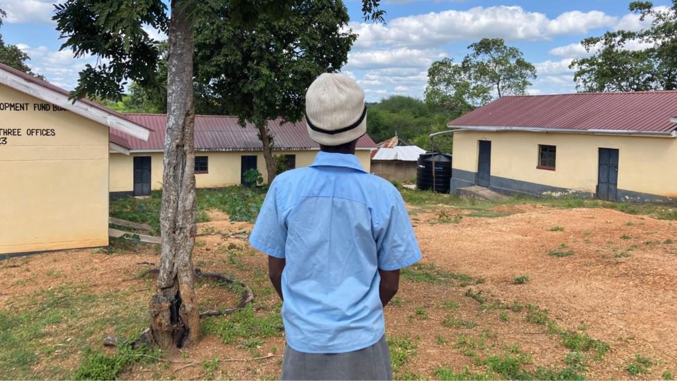 A student wearing a light blue short-sleeved school shirt, a grey skirt, and a cream-colored knit hat stands beneath a large tree in the courtyard of Manooni Primary School in Kenya