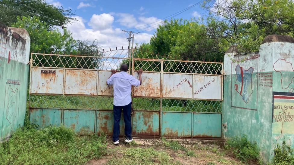 A man wearing a white shirt and black trouser stands at a large weathered metal gate of closed Sooma Primary School, peering through it into a green compound surrounded by trees