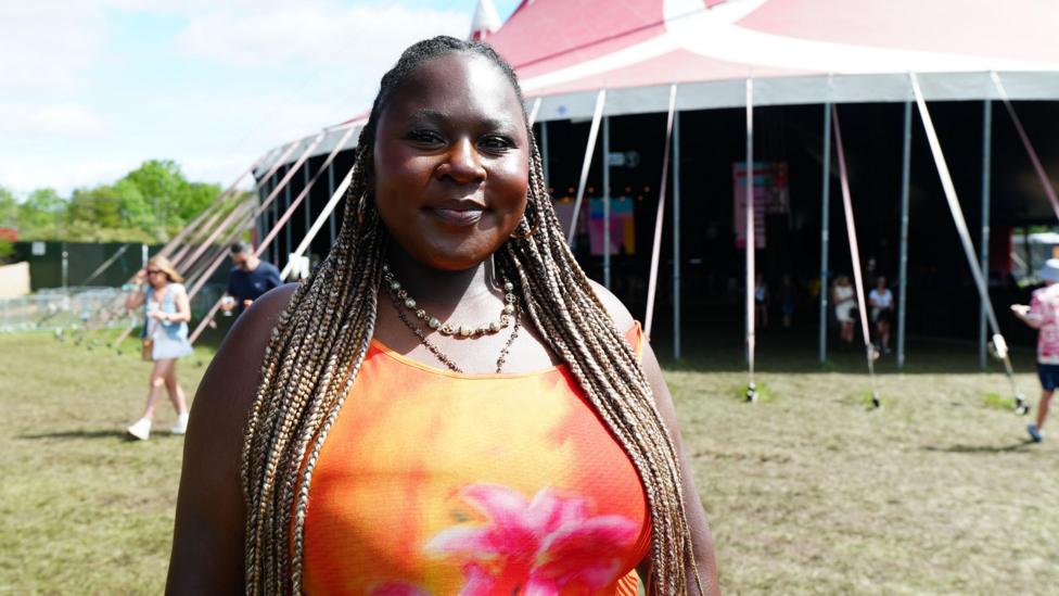 A woman with long hair styled in locs, wearing a blood-orange vest top stands outside a large tent at a music festival. People can be seen rushing about on the sun-drenched grass behind her.