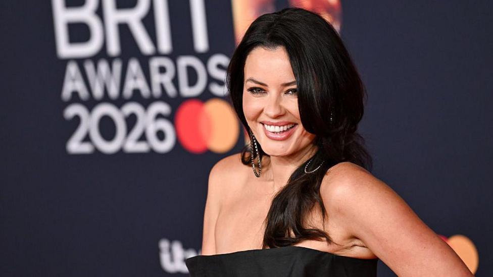 A woman with long black hair is smiling while wearing a black dress. She appears to be on a red carpet at the BRIT awards.