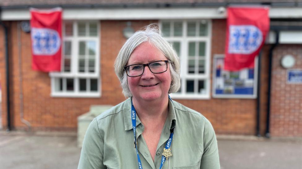 A woman with short grey hair, wearing black-rimmed glasses and a green shirt, smiling in front of a school building. At either side of her there is an England football badge flag. 
