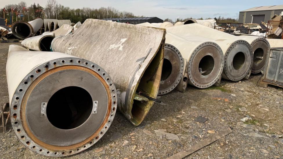 A row of disused wind turbine blades lie in an industrial yard. The white blades are rounded and hollow at one end but taper off into a flat blade in the distance. There is a warehouse and trees in the distance.