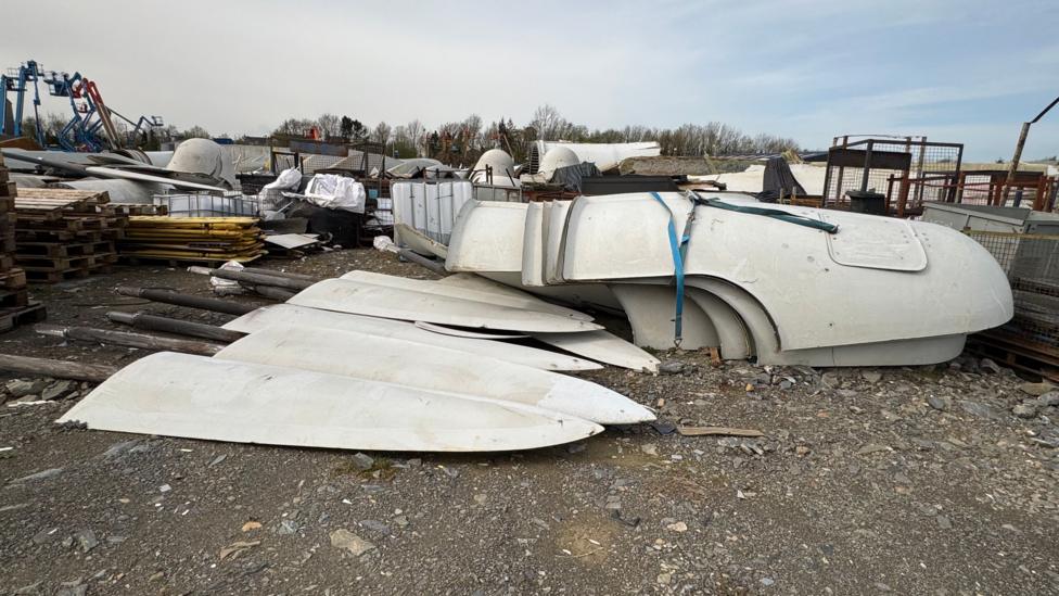Large piles of disused turbine waste is piled up in an industrial yard. It includes at least six large white tips of turbine blades.