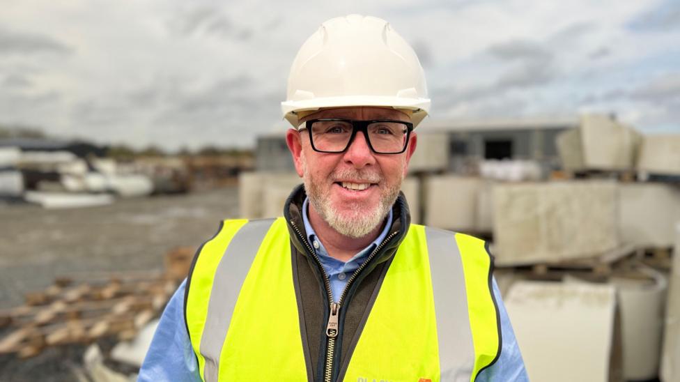 Kieran Kelly, a man with black square-framed glasses and a grey beard, smiles at the camera in an industrial yard. He is wearing a white helmet, a yellow high-viz vest, a blue shirt and a brown body warmer.