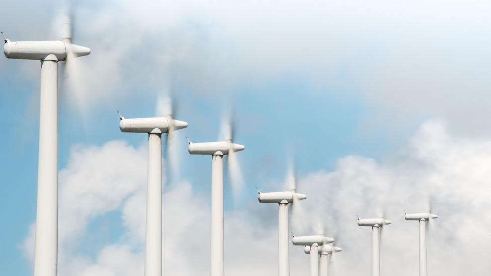 A row of white wind turbines rotating, pictured in front of a blue sky with fluffy white clouds.