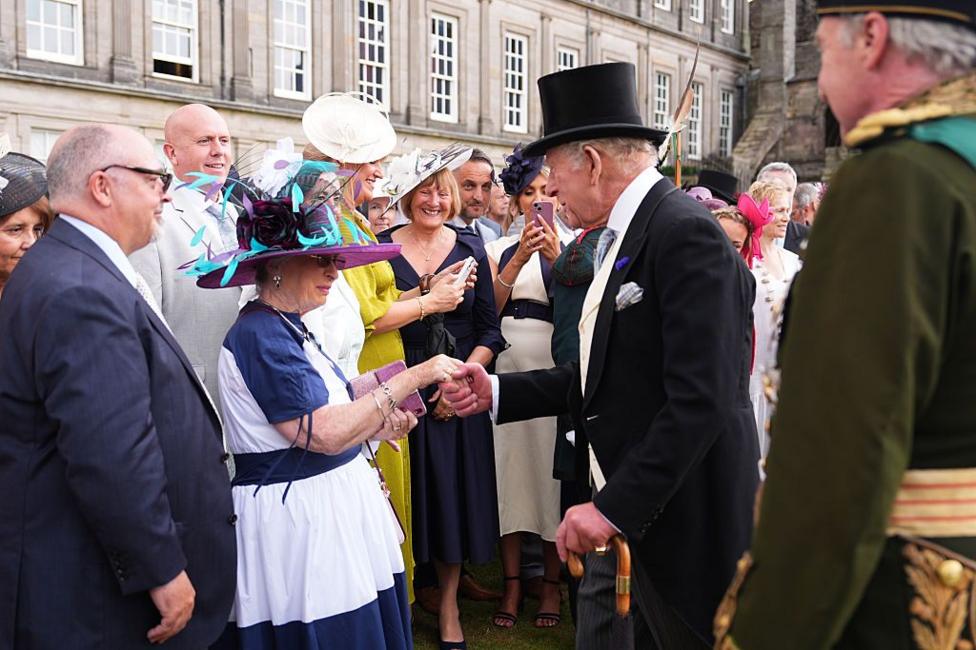 A crowd of people outside Holyroodhouse meet the King. He is smiling and holding the hand of a woman in a formal dress. The King is in a morning suit and top hat.