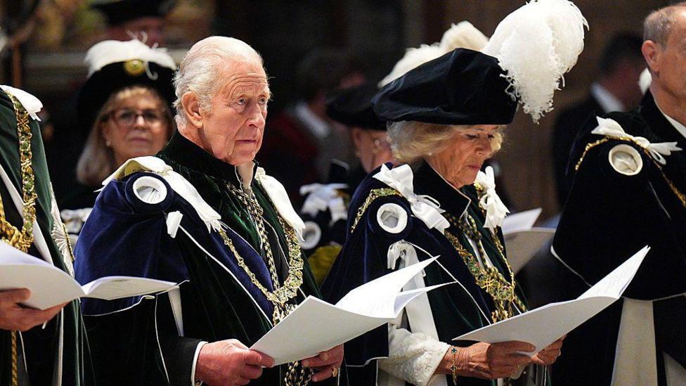 Queen Camilla and King Charles III attend the Thistle Service at St Giles' Cathedral. They are in ceremonial robes and are singing from sheets.