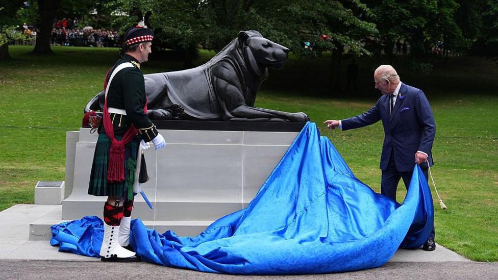The King and a soldier in full ceremonial dress, unveil a statue of a lion.
