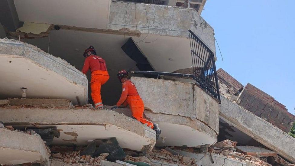 Two firefighters in orange uniforms climbing the rubble of a damaged building.