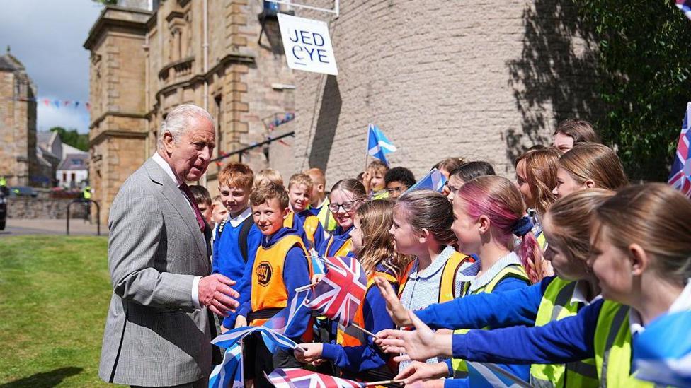 The King meeting local children. They are waving flags and looking happy and he is chatting away to them. Jedburgh Abbey can be seen in the background.