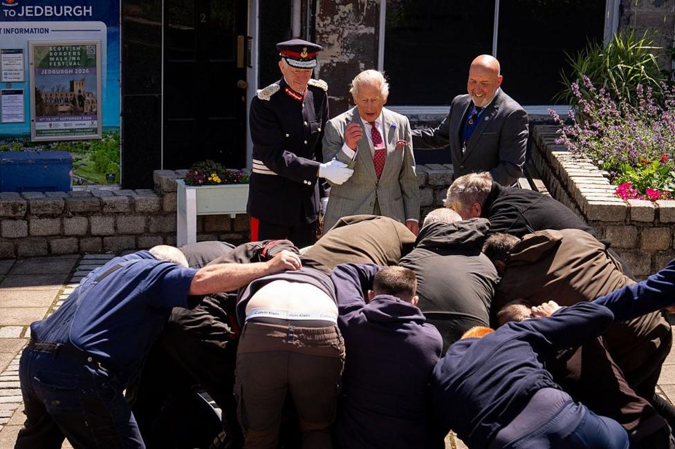 King Charles III is held back by a man in uniform and a man with a bald head as a group of men grapple for a ball in front of him