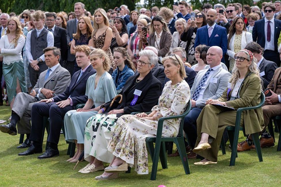 A group of people including Sophie, Duchess of Edinburgh, and Lady Louise Mountbatten-Windsor sit with other VIPs in the gardens of Holyroodhouse. There are many young people in the background.