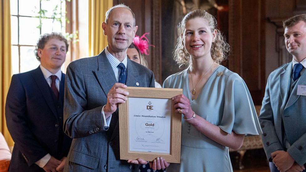 Prince Edward, Duke of Edinburgh presents his daughter Lady Louise Mountbatten-Windsor with the Duke of Edinburgh's Award Gold Award at Palace of Holyroodhouse. They are dressed formally and standing side-by-side, smiling. The award is a document in a wooden frame.