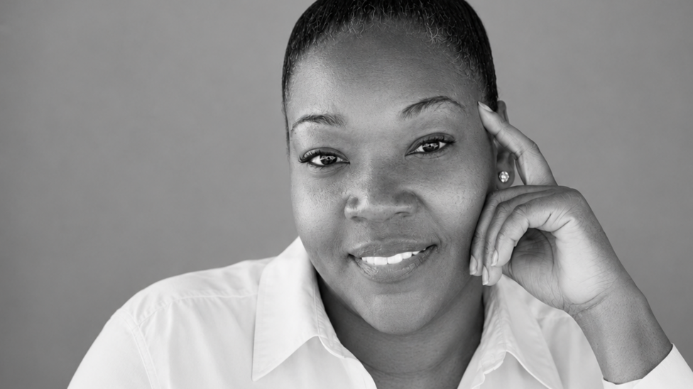 A black and white photo of a woman in a white blouse smiling into the camera. She has her hair drawn back off her face.
