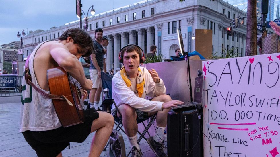 A man playing a guitar kneels next to a man sat in a chair alongside a sign that reads: