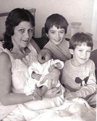 A woman in a hospital bed holdin a baby with two other young boys at the bedside.