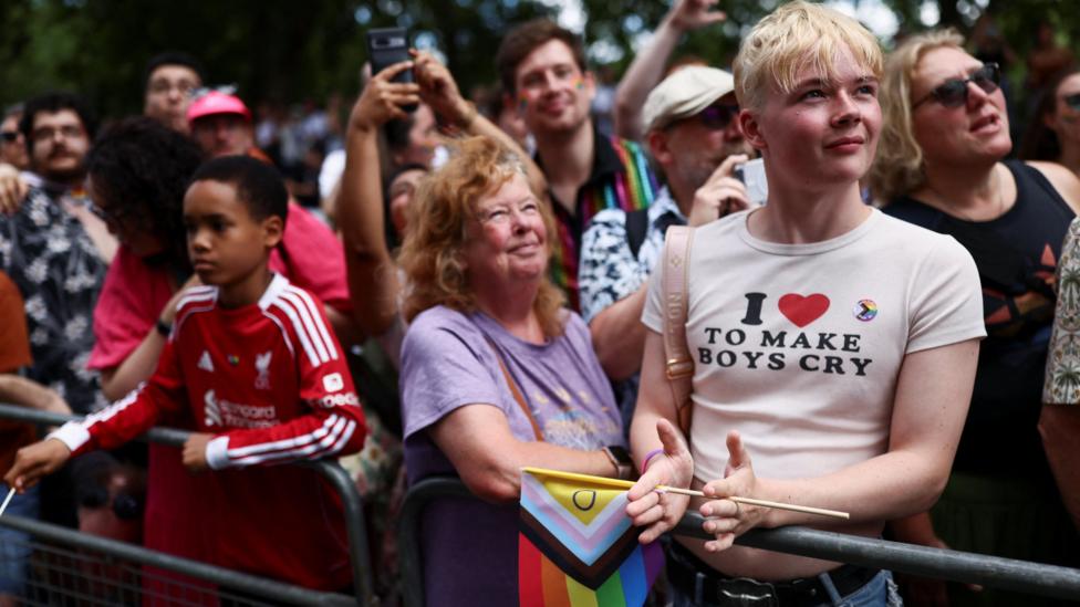 A crowd of spectators watching the parade from behind a metal barrier. In the foreground, a young person with short blonde hair claps while wearing a white cropped t-shirt that reads