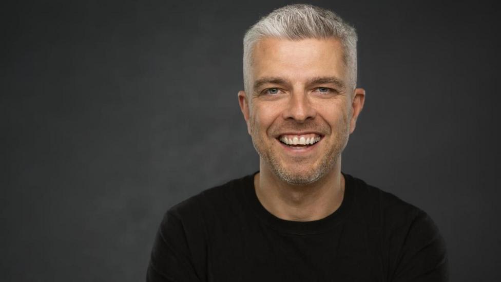 Peter has a full head of short grey hair, lifted at the front. He is wearing a black t-shirt and smiling.