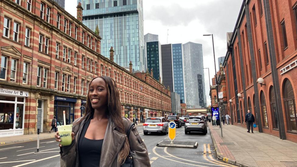 Ruwaydah in the foreground holding a clear plastic cup with a green drink and lid, standing in the middle of a city street. Behind, a traffic island with a yellow and blue directional bollard separates lanes of cars. Pavements line both sides with pedestrians and shopfronts, including signs reading “Millesime Art” and “Goodwin Fish”. Red-brick buildings flank the street, with modern glass high-rise towers in the background under overcast skies.