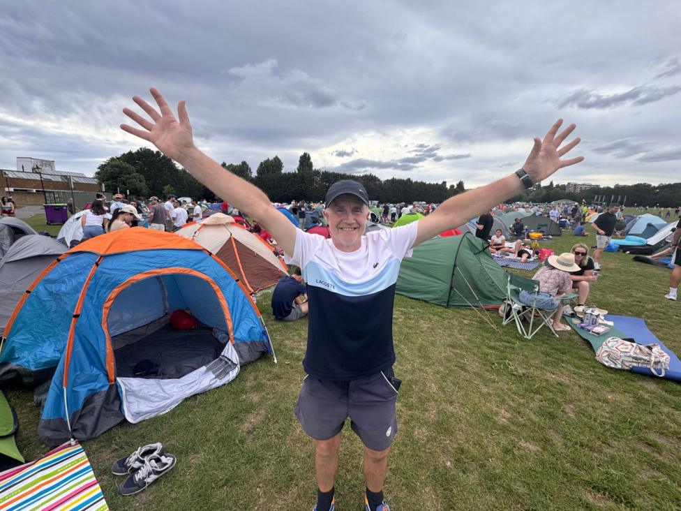 Geoff is holding a flag alongside a row of tents in the camping area of the queue