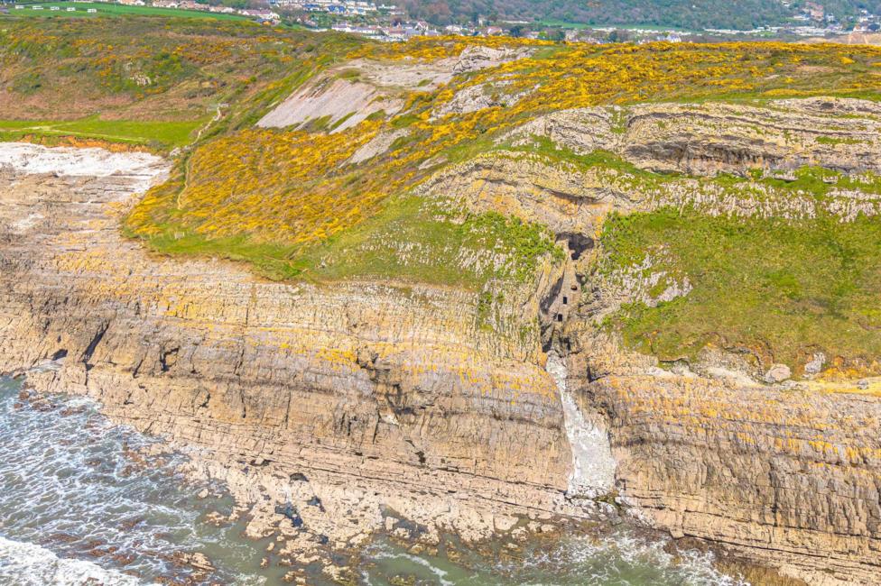 A high, drone-style view of a rugged coastal cliff layered with pale rock and streaks of yellow flowering vegetation. In the centre, a narrow gorge cuts down through the cliff, revealing a small, vertical stone structure embedded within the rock face. Below, waves break against scattered rocks along the shoreline. Above the cliffs, a grassy plateau stretches toward a nearby village with rows of houses and roads visible in the distance.
