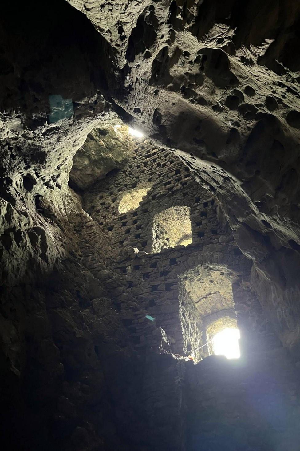 Interior view of a cave with a curved, textured sides with small holes. A vertical brick-wall inside, with narrow openings letting in bright daylight that contrasts sharply against the dark, enclosed space.