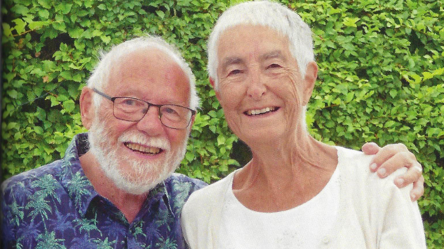 Malcolm Ridge stood on the left grinning, he has white hair and a white beard and mustache. He is wearing black framed glassed and a purple and green palm tree patterned shirt. He has his arm around Ruth who also has white hair and is smiling at the camera. She is wearing a white top.