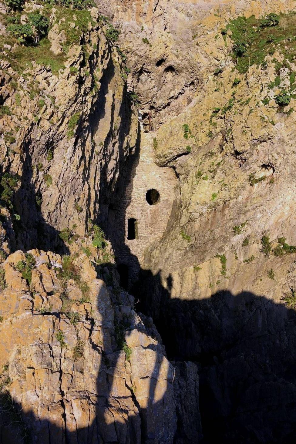 Stone structure with small round and rectangular openings built into a steep, rugged cliff face. Sunlight highlights the rough beige rock, while deep shadows fall into the narrow gorge below and sparse greenery clings to the rock surface.