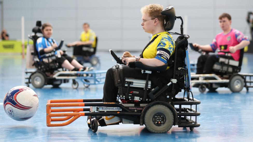 Picture of Dylan Kelsall during the match. He is sat in his wheelchair, with the ball ahead of him. He has blonde, curly hair and glasses.