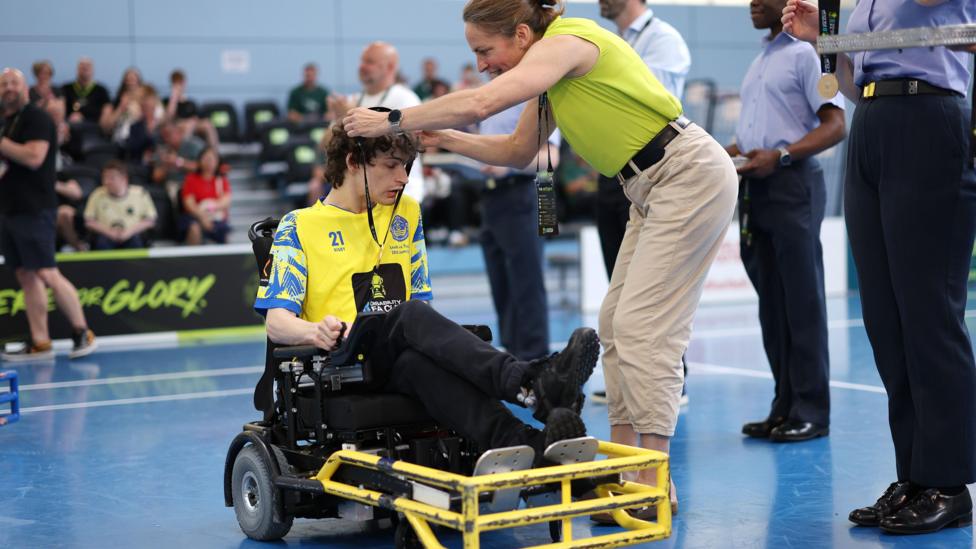 Dan gets his winners medal placed around his neck on court. He wears a yellow shirt. He has dark hair and is sat in his wheelchair. Several fans are pictured, sat, in the background.