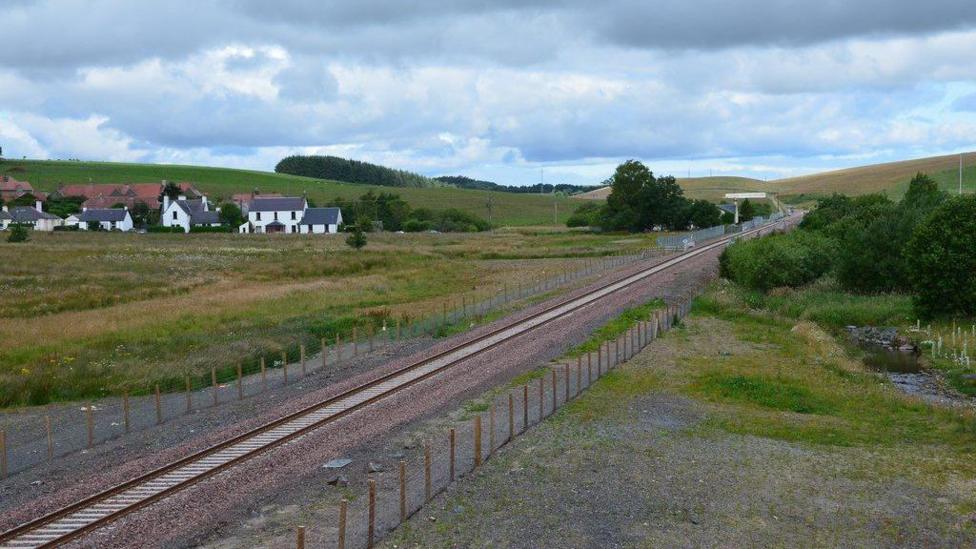 A railway line cuts through the Borders countryside under an ominous sky with a cluster of white houses to the left