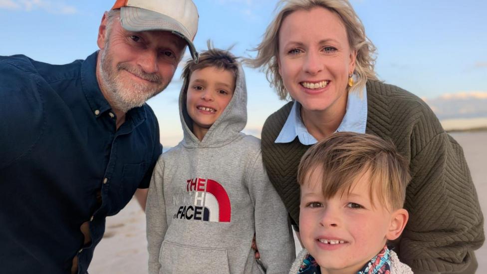 A family group with a man with a baseball cap on and a beard and a women with wavy blonde hair and two boys smiling into the camera. It looks like they are on a beach somewhere.