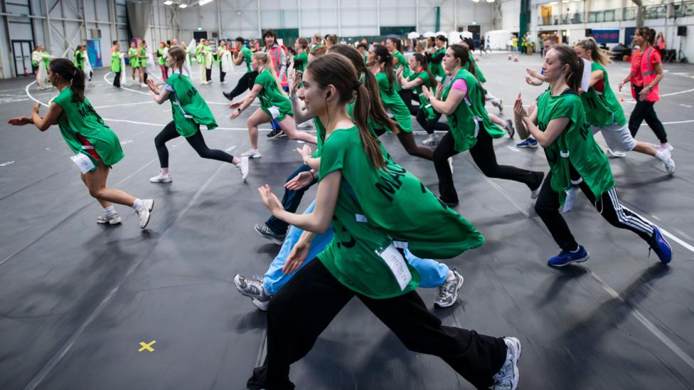 A group of dancers, mostly wearing green bibs, practice a routine inside a sports hall