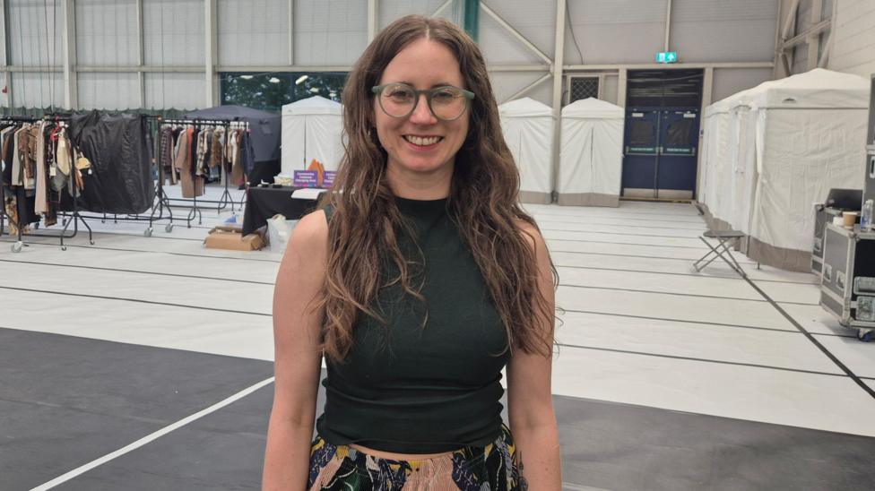 Roxana Cole - a woman with long brown hair and glasses, wearing a green top, standing in a large sports centre hall. Racks of costumes and clothing are behind her.