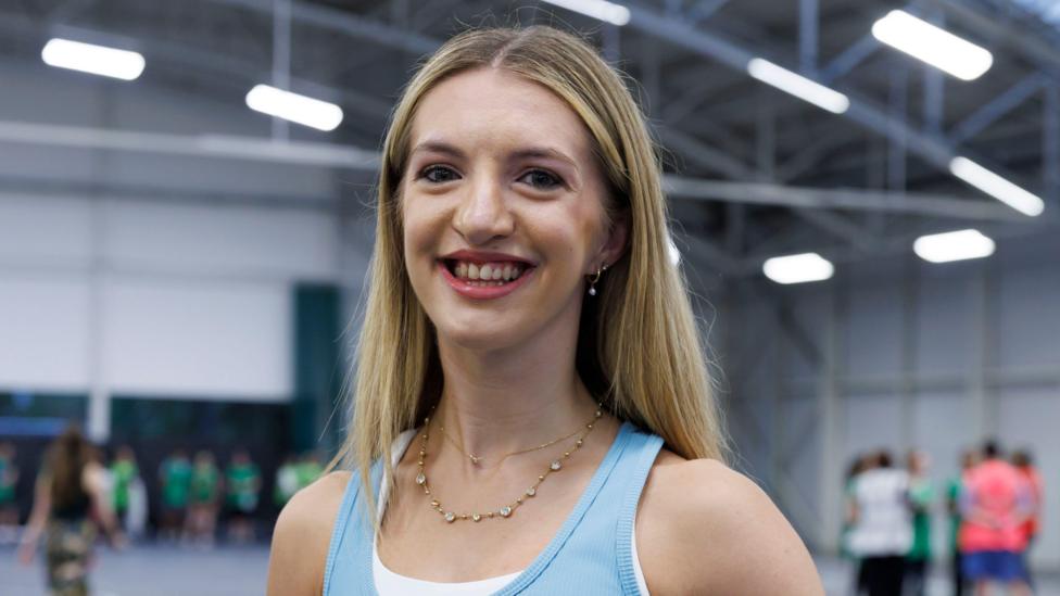 Kira Ewing - a woman with blond hair and a blue top, smiling for the camera during a dance rehearsal inside a sports hall.