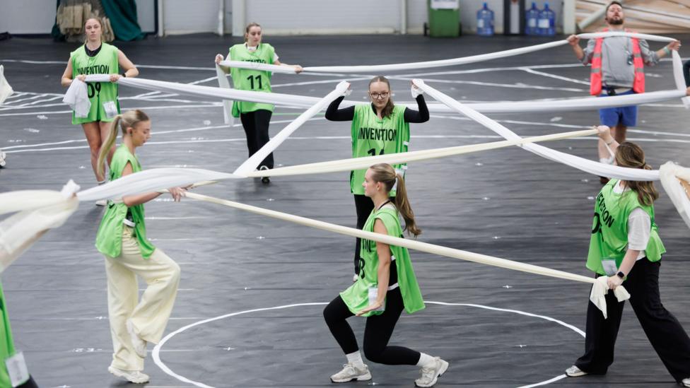 A group of dancers wearing light green bibs, holding fabrics as they practice a dance routine