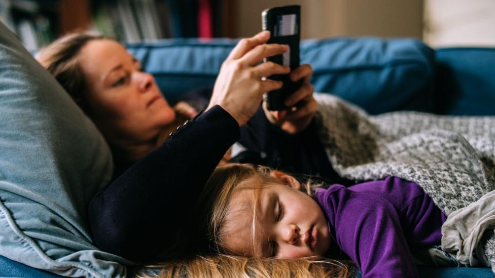 A young girl in a purple sweatshirt lies next to a woman in a black jumper on a couch. Both are covered in blankets
