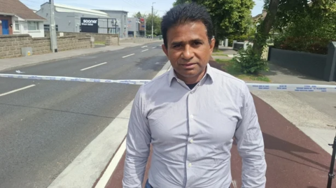 Thomas Joseph. He has dark hair, wearing a light coloured shirt. He is standing in front of a police cordon on a residential street.