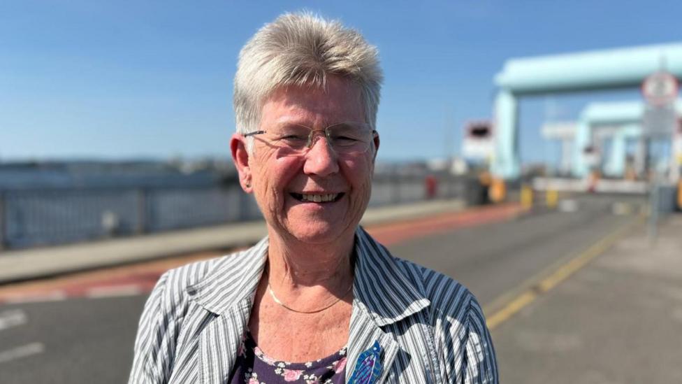 The image shows Jane Hutt - a middle-aged woman standing on the barrage. It it is a sunny day. She is wearing glasses and a stripey jacket.
